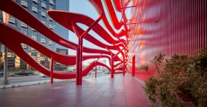 View from ground level showing the play on light between the dual steel ribbons and the red-painted corrugated aluminum rain-screen which acts as a backdrop. Each ribbon curve combines front and bottom surfaces in brushed stainless steel, while the back and top surfaces are made of textured aluminium coated with "hot-rod red" paint. The Petersen Museum. Photography by Tex Jernigan